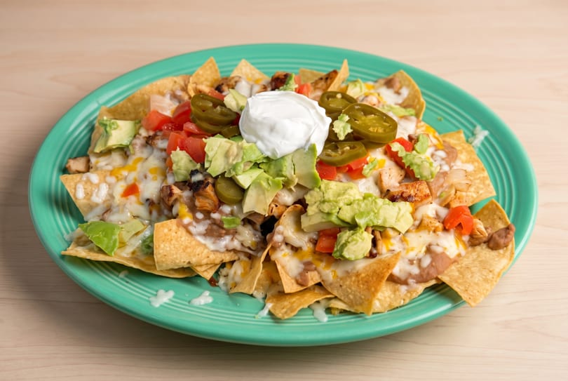 Nachos with melted cheese, chicken, beans, avocado, tomatoes, jalapeños, and sour cream on a teal plate.