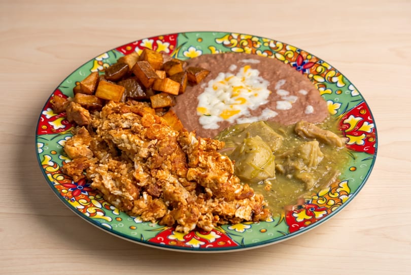 A colorful plate of scrambled eggs, refried beans with cheese, green chili, and roasted potatoes.