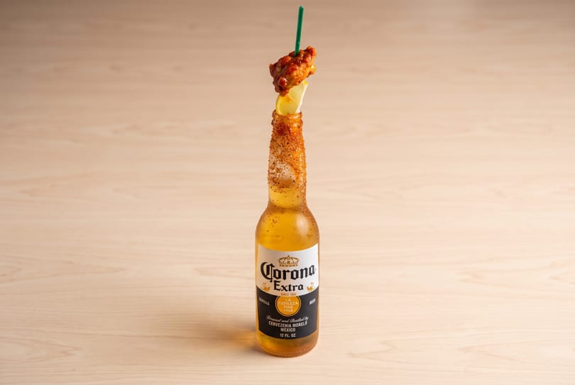 Corona Extra bottle with spiced rim, lemon, chicken wing, and green straw on a wooden table.
