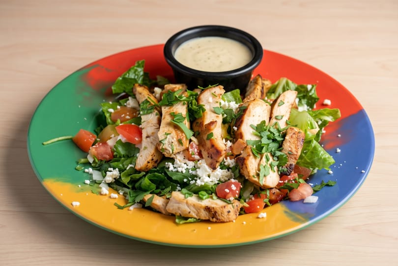 Grilled chicken salad with fresh greens, diced tomatoes, feta cheese, and dressing on a colorful plate.