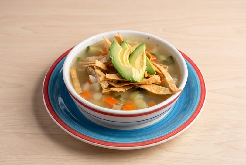 Tortilla soup with avocado slices and tortilla strips in a striped bowl on a wooden table.